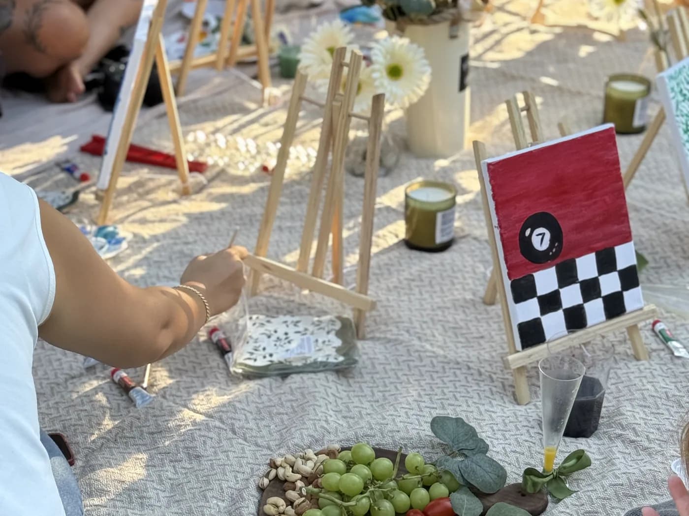 Small painted canvases, easels and snacks arranged on a picnic blanket