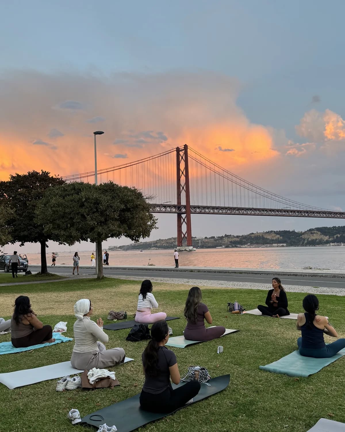 Women seated on yoga mats at sunset beside the river and Lisbon bridge