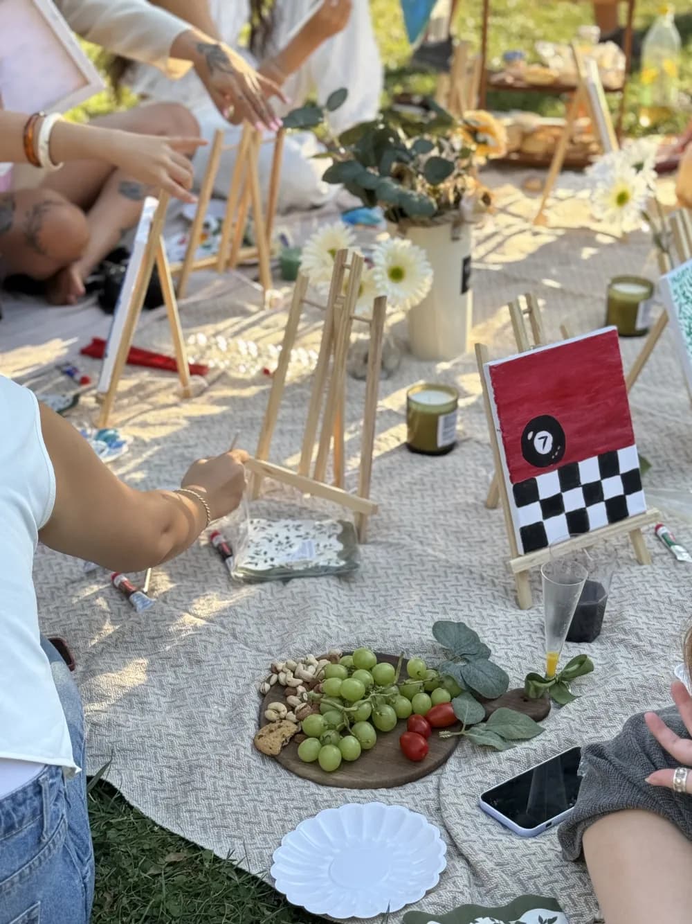 Small painted canvases, easels and snacks arranged on a picnic blanket